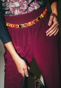 Pantaloni bordeaux con una cintura in vita testurizzata e una cintura a catena dorata con pietre ovali rosse, nere e arancioni, tenuti insieme da una borsa nera.