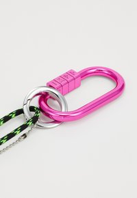 Rózsaszín fém karabiner zárkombinációval, ezüst színű körgyűrű, és fekete-zöld fonott pánt, amely ezüst láncon keresztül van rögzítve.