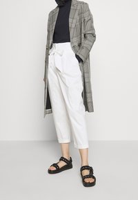 Geruite lange jas, zwarte geribbelde top, hoge witte broek met strikdetail en zwarte sandalen met ronde accenten.
