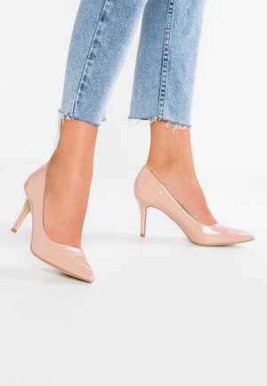 Jambes dans un jean léger bleu déchiré portant des escarpins beiges à bout pointu sur un fond blanc.