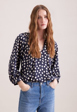 Kobieta o długich falujących włosach, ubrana w granatową bluzkę w białe kropki i niebieskie dżinsy, z rękami w kieszeniach, stojąca na jednolitym tle.