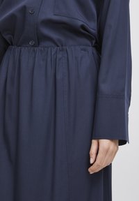 Robe bleu marine avec un devant boutonné, une taille élastique et des manches longues. Le matériau semble lisse avec un léger éclat. Détail de poche simple.