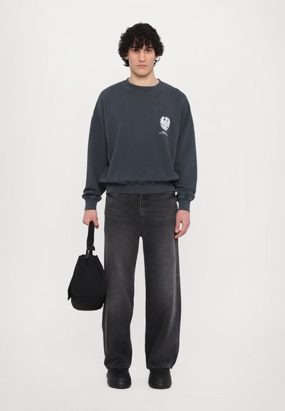 Szary sweatshirt z grafiką na klatce piersiowej, w połączeniu z luźnymi, ciemnymi dżinsami i czarnymi butami. Trzyma małą czarną torbę.