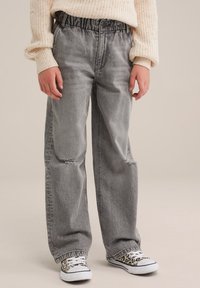 Šedé džínsy s širokými nohavicemi a elastickým pasem, s poškozením v oblasti kolen. V kombinaci se sneakerami s leopardím potiskem a pleteným svetrem.