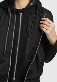 Czarna bomberka z kapturem, z metalowymi suwakami, bocznymi kieszeniami oraz teksturowanym materiałem. Wnętrze jest podszewkowane i dopasowane.