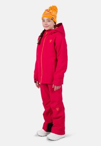 Rosa Skijacke und Hose mit gelben Akzenten und Logo. Die Jacke verfügt über eine Kapuze, einen Reißverschluss und Taschen. Weiße Stiefel vervollständigen das Outfit.
