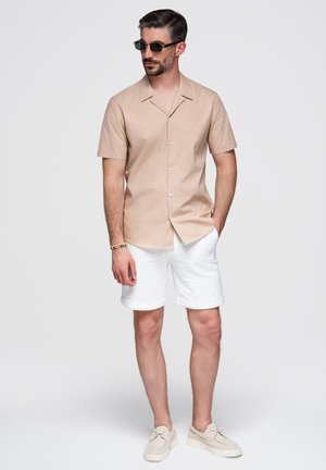 Mann mit Sonnenbrille, beigem, kurzärmeligem Hemd mit Knöpfen, weißen, hochgekrempelten Shorts und beigen Schuhen steht vor einem schlichten weißen Hintergrund.