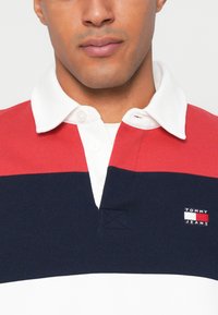 Rood, marineblauw en witte gestreepte poloshirt met een witte kraag, met een klein "TOMMY JEANS" logo op de borst en knoopsluiting.