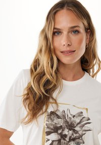 Bílá bavlněná tričko s černobílou květinovou grafikou v zlatém rámci. Krátké rukávy a kulatý výstřih. Hladká textura.
