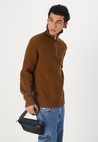 Brauner ribbeldesign Halzip-Pullover mit langen Ärmeln, kombiniert mit blauen Jeans. Das Model hält eine kleine schwarze Tasche mit einem Gurt.