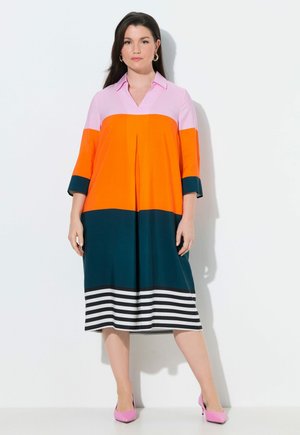 Žena stojící a nosící šaty s bloky barev, růžovým límečkem, oranžovou střední částí, tmavě zelenou spodní sekcí, pruhovaným lemem a růžovými lodičkami na podpatku.