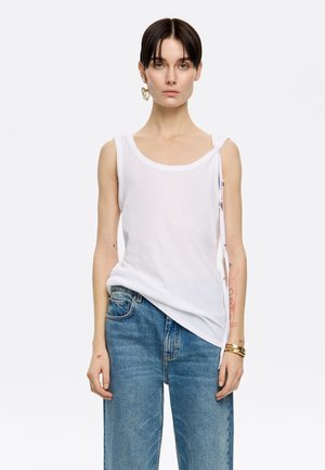 Top in cotone bianco senza maniche con spalline larghe, dotato di scollo rotondo e dettagli in pizzo sui lati. Abbinato a jeans blu larghi.