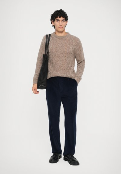 Hombre vestido con un suéter de punto beige, pantalones de pana azul marino, zapatos negros y llevando un bolso negro de hombro, contra un fondo blanco liso.
