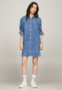 Vestido camisero de mezclilla azul con cierre de botones, dos bolsillos en el pecho, mangas enrolladas y una silueta recta. Se lleva con zapatillas blancas y calcetines.