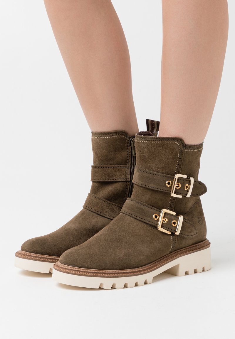 Bottines marron en daim avec fermeture éclair latérale, doubles boucles dorées et semelles blanches en caoutchouc épaisses, portées sur des jambes nues.