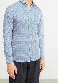 Camisa de manga larga azul con estampado, que presenta un cuello abotonado, un diseño geométrico y un corte ajustado. La tela parece suave y ligera.