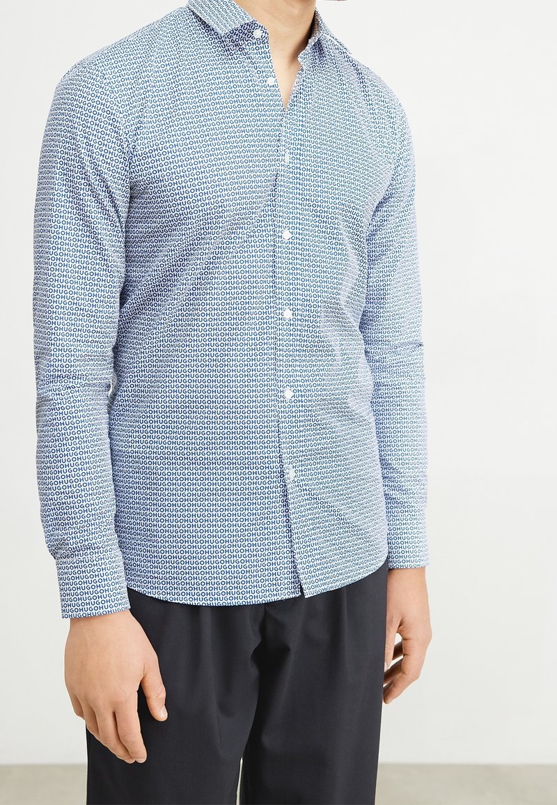 Camisa de manga larga azul con estampado, que presenta un cuello abotonado, un diseño geométrico y un corte ajustado. La tela parece suave y ligera.