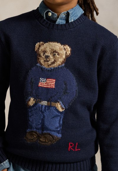 Enfant portant un pull en maille bleu marine avec un ours en peluche portant un jean, une ceinture, un pull avec un drapeau américain, et des chaussures marron tricotées à l'avant.