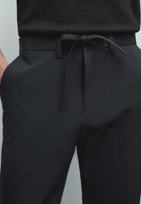 Gros plan sur un pantalon noir avec un cordon de serrage noué en forme de nœud et une main reposant dans la poche gauche sur un fond uni.