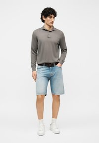 Άνδρας που φορά γκρι πόλο με μακριά μανίκια, ανοιχτόχρωμα μπλε τζιν σορτς με μαύρη ζώνη, λευκές κάλτσες και λευκά αθλητικά παπούτσια, στέκεται μπροστά σε λευκό φόντο.