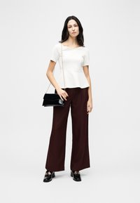 Femme portant un haut péplum blanc à manches courtes, un pantalon large bordeaux, des mocassins noirs à talons et tenant un sac à main en cuir verni noir avec une chaîne dorée.