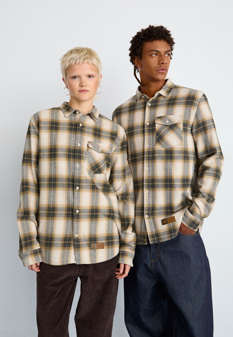 Dwie osoby noszą beżowe i w kratę koszule z guzikami, z ciemnozielonymi i żółtymi akcentami. Koszule mają kieszenie na piersiach i teksturowany materiał.