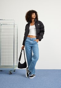 Svart pufferjakke, hvit crop tank topp, blå vide jeans, svart skulderveske, svarte og hvite sneakers. Enkel bakgrunn med metallstativ.