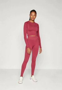 Žena stoji u bordo dugoj majici s kratkim rukavima i odgovarajućim tajicama visokog struka uz bijele tenisice na jednobojnoj pozadini.