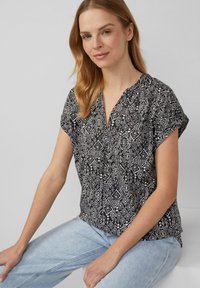 Schwarz-weiße gemusterte Kurzarmbluse mit V-Ausschnitt, die geometrische Designs und einen leichten Stoff aufweist. Kombiniert mit hellblauen Jeans.