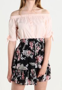 Helles rosa Off-Shoulder-Bluse mit kurzen Puffärmeln und Knopfdetails, kombiniert mit einem schwarzen Rock mit floralem Druck in Stufen.