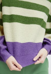Csíkozott pulóver krémszínű és zöld vízszintes csíkokkal, élénk lila alsó szegéllyel, valamint texturált kötött anyagból készült, felgöndörített ujjakkal.