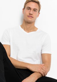 T-shirt bianco a maniche corte e scollo a V, realizzato in tessuto leggero, indossato con pantaloni neri. Texture liscia, design casual, dettaglio di scollo visibile.