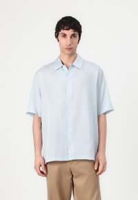Camicetta leggera azzurro chiaro a maniche corte con vestibilità rilassata, colletto button-down e tessuto texture, indossata sopra pantaloni beige.