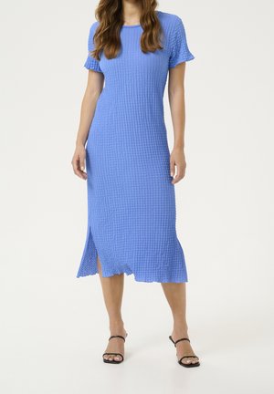Woman wearing a textured blue midi dress with short sleeves and side slit, paired with black open-toe heels against a plain background.