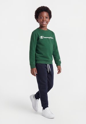 Dreng smilende og gående iført en grøn Champion sweatshirt, marineblå joggingbukser med hvide snore og hvide sneakers mod en hvid baggrund.