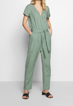 Zielony lniany kombinezon z dekoltem w serek, krótkimi rękawami, paskiem w talii i luźnym krojem. Posiada boczne kieszenie i zwężane nogawki.