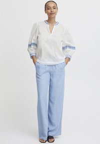 Blouse blanche avec broderie bleue sur les manches, dotée d'un col en V et d'une coupe ample. Associée à un pantalon large bleu clair et des sandales noires.