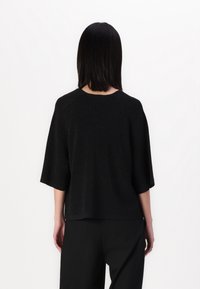 Femme aux cheveux noirs lisses portant un pull noir ample à manches courtes et un pantalon noir, debout face à un mur blanc.