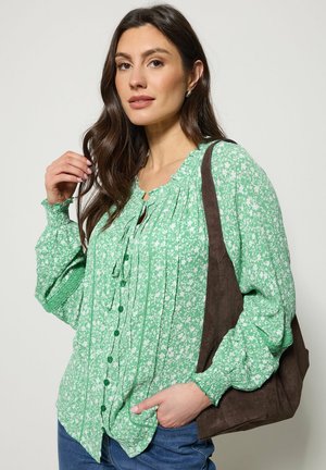 Frau trägt eine grüne, langärmelige Bluse mit Blumenmuster, blaue Jeans, hält eine braune Wildleder-Schultertasche vor einem schlichten Hintergrund.