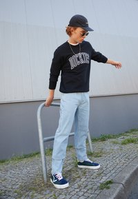 Black sweatshirt with "DICKIES" logo, light blue denim pants, and blue suede sneakers. Accessories include a navy cap and round sunglasses.