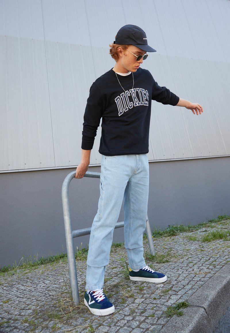 Black sweatshirt with "DICKIES" logo, light blue denim pants, and blue suede sneakers. Accessories include a navy cap and round sunglasses.