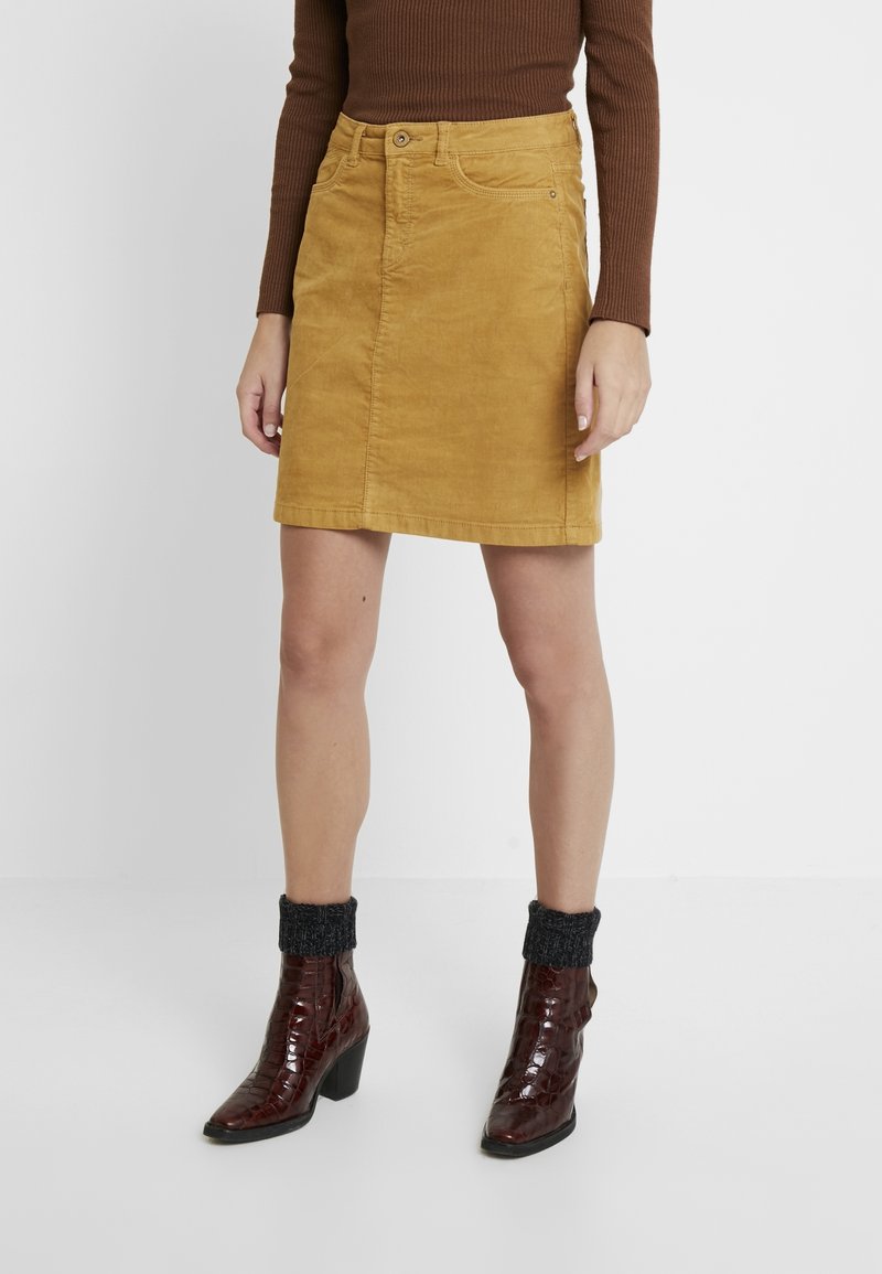 Femme portant une jupe en velours côtelé jaune moutarde, un haut marron à manches longues, des chaussettes gris foncé et des bottines à talons marron foncé, debout sur un fond blanc.