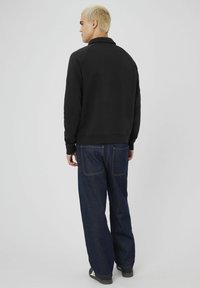 Jeune personne avec des cheveux blonds courts portant un sweat à capuche noir, un jean ample bleu foncé et des chaussures noires, se tenant de profil.