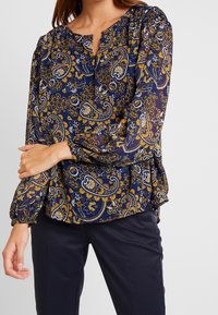 Blusa navy con un motivo floreale e paisley in giallo e bianco. Presenta un collo a V e maniche lunghe arricciate, realizzata in tessuto leggero.