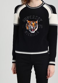 Pull noir en tricot avec des rayures blanches. Présente un graphisme de tigre et le texte « Superdry Michigan 1975 ». Poignets et ourlet côtelés.