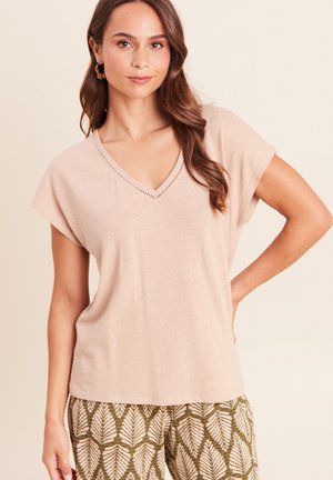 Femme aux longs cheveux bruns porte un haut beige à manches courtes en V et un pantalon à motifs avec de grands dessins de feuilles, posant avec une main sur la hanche.