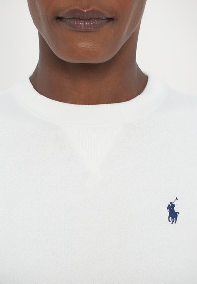 Gros plan sur une personne portant un sweat-shirt blanc à col rond avec un petit logo de joueur de polo bleu marine sur la poitrine gauche.