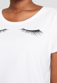 Camiseta de algodón blanca con cuello redondo y mangas cortas, decorada con gráficos de pestañas negras en la parte delantera.