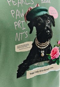 Zielony materiał z czarnym psem w zielonym berecie i perłowej naszyjniku, w towarzystwie kwiatowych akcentów oraz stylizowanego tekstu.