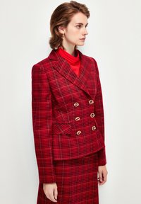 Röd rutigt dubbelknäppt kavaj med guldfärgad hårdvara, med en strukturerad design och textil med struktur, tillsammans med en röd polotröja.